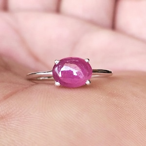 Puede incluir: Un anillo de plata con una gran gema rosa vibrante de forma ovalada. La gema está sujeta por cuatro pequeñas garras. La banda del anillo es delgada y pulida, creando un diseño simple pero elegante. El anillo se muestra en una mano.
