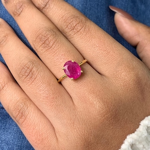 Op de afbeelding: Een gouden ring met een grote, ovaal geslepen, levendige roze-rode edelsteen in een zetting met vier pootjes. De ringband is dun, gepolijst en van goudkleurig metaal, wat een klassiek ontwerp vormt dat de verzadigde kleur benadrukt.