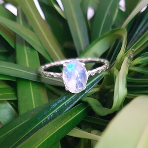 Puede incluir: Anillo de plata con una gran piedra lunar ovalada. La piedra lunar tiene un aspecto lechoso e iridiscente con destellos azules y verdes. La banda tiene un diseño texturizado y martillado, sobre un fondo de hojas verdes.