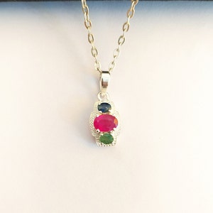 Peut inclure: Un collier en argent avec un pendentif composé de trois pierres précieuses ovales : un rubis rouge au centre, un saphir bleu au-dessus et une émeraude verte en dessous.