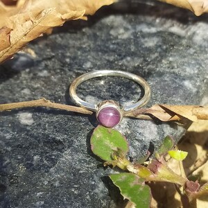 May include: A silver ring with a round, purple gemstone set in the centre. The ring is on a grey rock with green leaves.