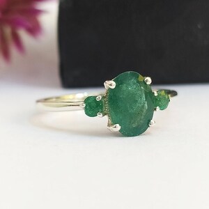 Puede incluir: Un anillo de plata con una gran piedra preciosa verde de corte ovalado en el centro, flanqueada por dos piedras verdes más pequeñas de corte redondo a cada lado. Las piedras están sujetas por garras de plata sobre un aro liso y pulido.