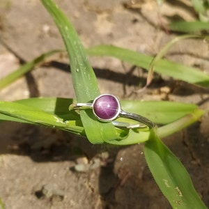 May include: A silver ring featuring a purple star sapphire gemstone. The ring is resting on a green leaf.
