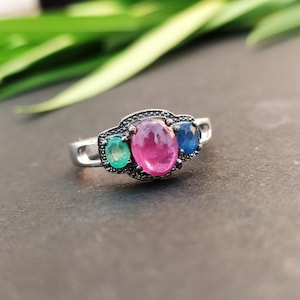 May include: A silver-toned ring featuring a central oval-shaped pink gemstone flanked by a smaller oval green gemstone on the left and a smaller oval blue gemstone on the right, set within a textured, darkened metal frame with a polished band.