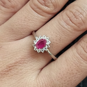 Könnte beinhalten: Ein silberner Ring mit einem ovalen, leuchtend roten Rubin, umgeben von kleineren, klaren Edelsteinen. Der Ring hat ein zartes, poliertes Band. Ein klassisches und elegantes Design.