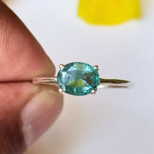 May include: A silver ring featuring a teal oval gemstone. The gemstone is set with four prongs and has a faceted surface. The ring is displayed against a soft, blurred background.