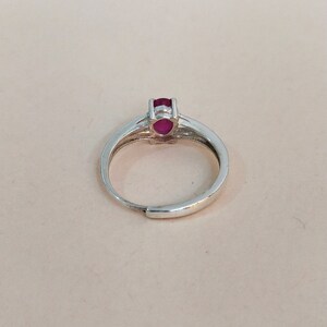 Puede incluir: Un anillo de tono plateado con una peque&ntilde;a piedra preciosa ovalada de color rojo rosado intenso en un engaste de garras. La banda es lisa y met&aacute;lica con una abertura ajustable en la parte inferior, mostrando un dise&ntilde;o simple y funcional.