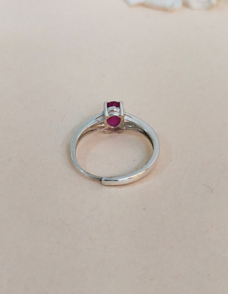 Puede incluir: Un anillo de tono plateado con una peque&ntilde;a piedra preciosa ovalada de color rojo rosado intenso en un engaste de garras. La banda es lisa y met&aacute;lica con una abertura ajustable en la parte inferior, mostrando un dise&ntilde;o simple y funcional.