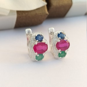 May include: A pair of silver hoop earrings with three gemstones. The earrings feature a large oval-shaped ruby, a smaller round sapphire, and a smaller oval-shaped emerald.