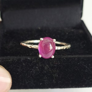 May include: A silver ring featuring a smooth, oval-shaped, vibrant pink cabochon gemstone set in a four-prong setting. The ring band has a textured, hammered finish, providing a distinct contrast to the polished surface of the central stone.