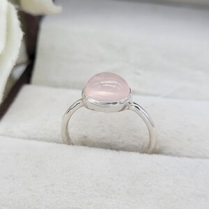 May include: A silver ring featuring a large, oval-shaped rose quartz gemstone. The pale pink stone is set in a simple, polished silver band. The ring is displayed on a white surface, highlighting its delicate color and design.