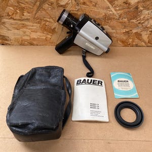 Op de afbeelding: Een vintage Bauer C4 Super 8 filmcamera met een zwarte leren draagtas, handleiding en lensdop. De camera is zilver en zwart met een zwarte lens.