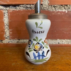 May include: White ceramic sugar dispenser with a silver metal spout and lid. The dispenser is decorated with colorful floral designs and the word "Sucre" in black script. The dispenser is on a wooden surface.