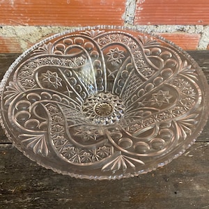 May include: Clear glass serving bowl with intricate star and floral patterns. The bowl has a scalloped edge and a central, raised base. The design features repeating shapes and textures, creating a vintage aesthetic.