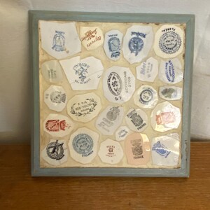 May include: A framed artwork composed of vintage porcelain fragments, each bearing stamps and designs. The fragments are set against a beige backdrop within a light blue wooden frame. Text includes "FRANCE" and "BENGAL".
