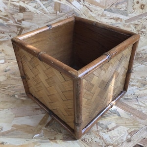 Puede incluir: Una jardinera cuadrada de madera con un diseño de bambú tejido. La jardinera tiene un acabado de madera natural y detalles de bambú en los bordes. El interior es de un tono de madera más oscuro.