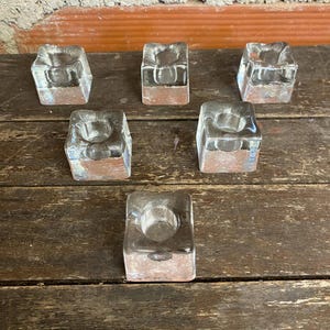 Puede incluir: Seis candeleros cuadrados de vidrio transparente dispuestos sobre una superficie de madera. Cada candelero tiene una base cuadrada y una hendidura circular para una vela. El vidrio transparente permite el paso de la luz.