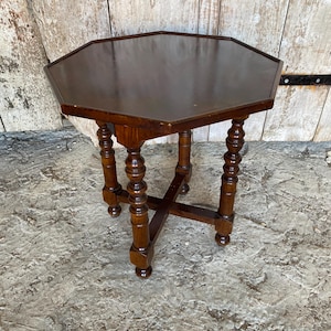 May include: A dark brown wooden side table with an octagonal tabletop. The table features four legs with a turned, spindle-style design, connected by a cross-shaped stretcher base that provides structural support to the legs.