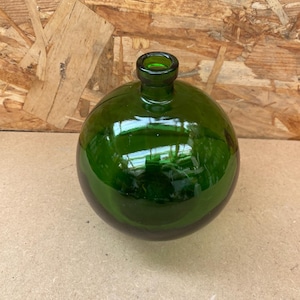 Puede incluir: Una botella redonda de vidrio verde oscuro con un cuello pequeño. La botella tiene una superficie lisa y reflectante, y el color verde es uniforme. La botella está sobre una superficie marrón claro.