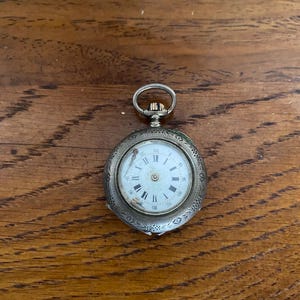 May include: A silver pocket watch with an oval-shaped case and a white face with black Roman numeral hour markers. The watch has a decorative floral design around the edge of the case.