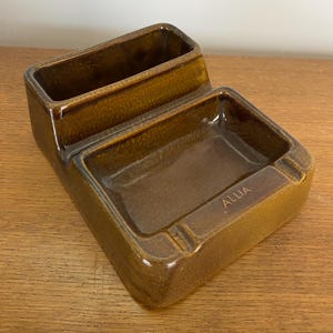 Peut inclure: Un cendrier en céramique brun avec un compartiment rectangulaire plus petit à côté. Le cendrier est marqué "ALLIA".