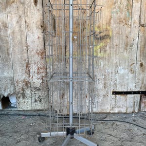 Könnte beinhalten: Ein drehbarer Präsentationsständer aus silberfarbenem Metall mit mehreren Drahtregalen. Der Ständer hat eine zentrale Stange und einen Sockel mit Rollen für eine einfache Bewegung. Der Hintergrund ist eine verwitterte Holzwand.