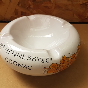以下が含まれることがあります： 白いセラミック灰皿。黒字で「JAS HENNESSY & CO COGNAC」と書かれています。灰皿は丸みを帯びた形で、タバコ用の3つのくぼみと、オレンジ色のブドウの装飾的な縁があります。