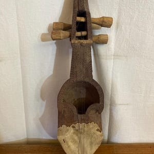 Può includere: Strumento musicale a corde in legno d'epoca, con corpo e manico intagliati. Presenta una cassa armonica cava, una membrana chiara e quattro piroli in legno. Il legno ha un aspetto scuro e invecchiato.