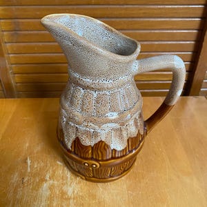 May include: A ceramic jug featuring a two-tone design with a speckled beige and white upper section and a dark brown, wood-grain textured lower section. The jug has a curved handle and a wide pouring spout with a distinct drip-like glaze finish.