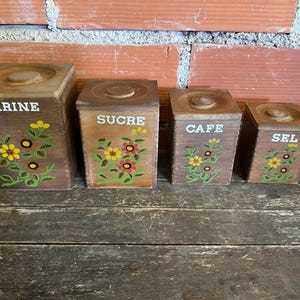 May include: Set of four wooden kitchen canisters with lids. Each canister is a different size and labeled with the words "FARINE", "SUCRE", "CAFE", and "SEL" in white lettering. Floral designs in yellow, red, and green adorn the front of each canister.