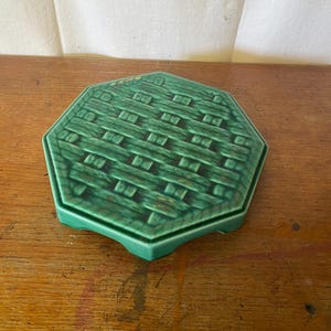 Puede incluir: Un salvamanteles de cerámica verde con un diseño de cesta tejida. El salvamanteles es hexagonal y tiene un acabado brillante. Está colocado sobre una superficie de madera.