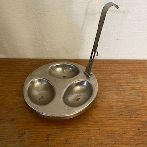 Puede incluir: Un escalfador de huevos vintage, de tono plateado, con tres copas de forma ovalada. Cuenta con un asa con un gancho para colgar. El escalfador está diseñado para cocinar tres huevos simultáneamente. El metal tiene un aspecto ligeramente envejecido.