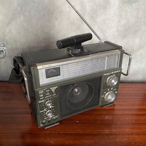 Puede incluir: Una radio Venture vintage de color verde y negro con una antena telescópica, un dial con números de frecuencia y varios botones para sintonizar, volumen y tono. La radio tiene un altavoz en la parte delantera y una correa de transporte.