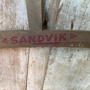 Puede incluir: Primer plano de una tabla de madera con el texto rojo "SANDVIK" y una flecha roja apuntando hacia la derecha. El texto "AGUSA SWEDEN" también es visible en letras rojas más pequeñas.