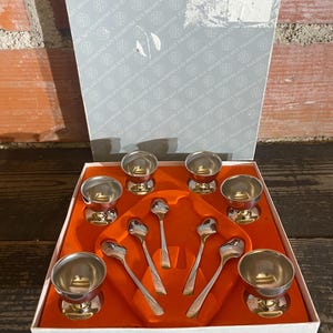 Puede incluir: Un juego de tazas de huevo y cucharas vintage, presentado en una caja con interior naranja. Las tazas de huevo son plateadas, con base de pedestal. Las cucharas son delgadas y plateadas. La caja tiene el texto "GUY DEGRENNE" impreso.