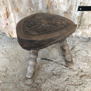 Könnte beinhalten: Ein rustikaler Holzhocker mit drei gedrehten Beinen und einer dreieckigen Sitzfläche. Die Sitzfläche hat ein geschnitztes Muster und die Beine sind in einem helleren Holzton gehalten.