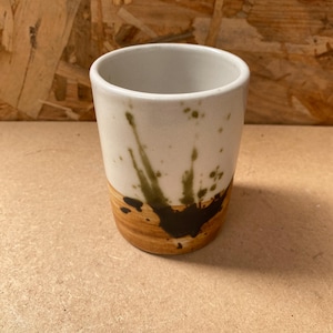 Puede incluir: Una taza de cerámica blanca con salpicaduras de pintura marrón y verde. La taza tiene un fondo redondeado y una abertura ancha.