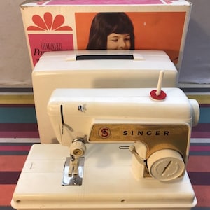 May include: A vintage Singer Golden Panoramic sewing machine in cream and gold. The machine features a gold Singer logo and a white spool pin. The original box is behind the machine, with a picture of a girl on the front.