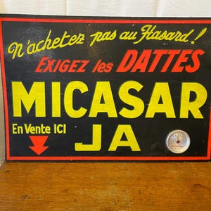 May include: Vintage wooden sign with black background and yellow and red lettering. The sign reads "N'achetez pas au Hasard! EXIGEZ les DATTES MICASAR JA En Vente ICI" with a small gauge at the bottom.