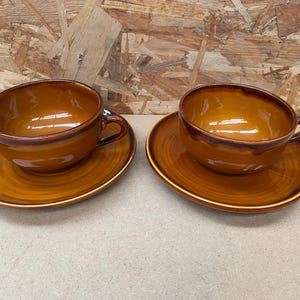 May include: Two brown ceramic teacups with matching saucers. The cups and saucers have a glossy finish and a swirling pattern.