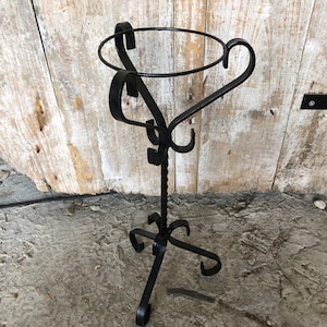 Puede incluir: Soporte para plantas de metal negro con parte superior redonda y tallo retorcido. El soporte tiene un diseño decorativo con volutas y curvas.