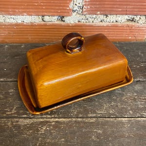 May include: A rectangular, ceramic butter dish with a lid and a handle. The dish and lid are a warm, glossy brown colour. The handle is a small, ring-shaped design. The dish sits on a matching rectangular base.