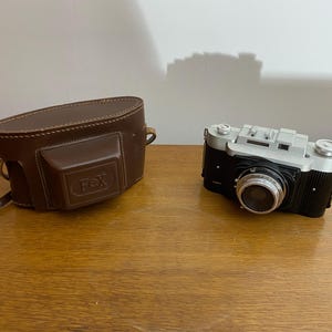 May include: A vintage silver and black camera with a brown leather case. The camera has a lens with a silver ring and the case has the word "FOX" embossed on it.