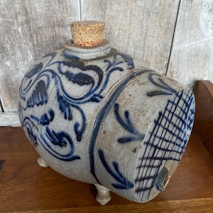 May include: A blue and white stoneware crock with a cork stopper and three legs. The crock is decorated with a blue and white floral design.