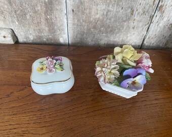 May include: Two porcelain trinket boxes with floral designs. One box is a square shape with a lid and the other is a flower shaped box with a bouquet of flowers.