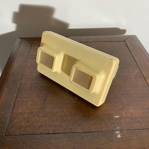 Peut inclure: Un ancien View-Master de couleur crème. L'appareil rectangulaire comporte deux trous pour les yeux et une forme rectangulaire. Le lecteur repose sur une table en bois brun foncé.
