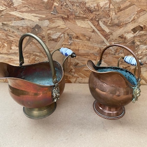 May include: Two antique copper coal scuttles with blue and white porcelain handles. The scuttles have a rounded shape and are made of polished copper.