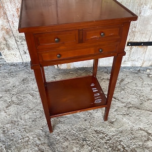 May include: A small, dark brown wooden side table with two drawers and a lower shelf. The table has a rectangular top and stands on four slender legs. The drawers have small, round knobs. The table is approximately 28 inches tall.