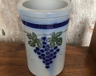 Peut inclure: Un pot en grès avec une finition bleu clair et blanche. Le pot présente une bande bleu foncé et un motif de grappe de raisin avec des feuilles vertes. Le pot est cylindrique avec une large ouverture.