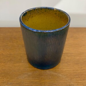Peut inclure: Un petit vase en céramique cylindrique avec un glaçage bleu et jaune. Le vase a une surface texturée et un bord légèrement évasé.